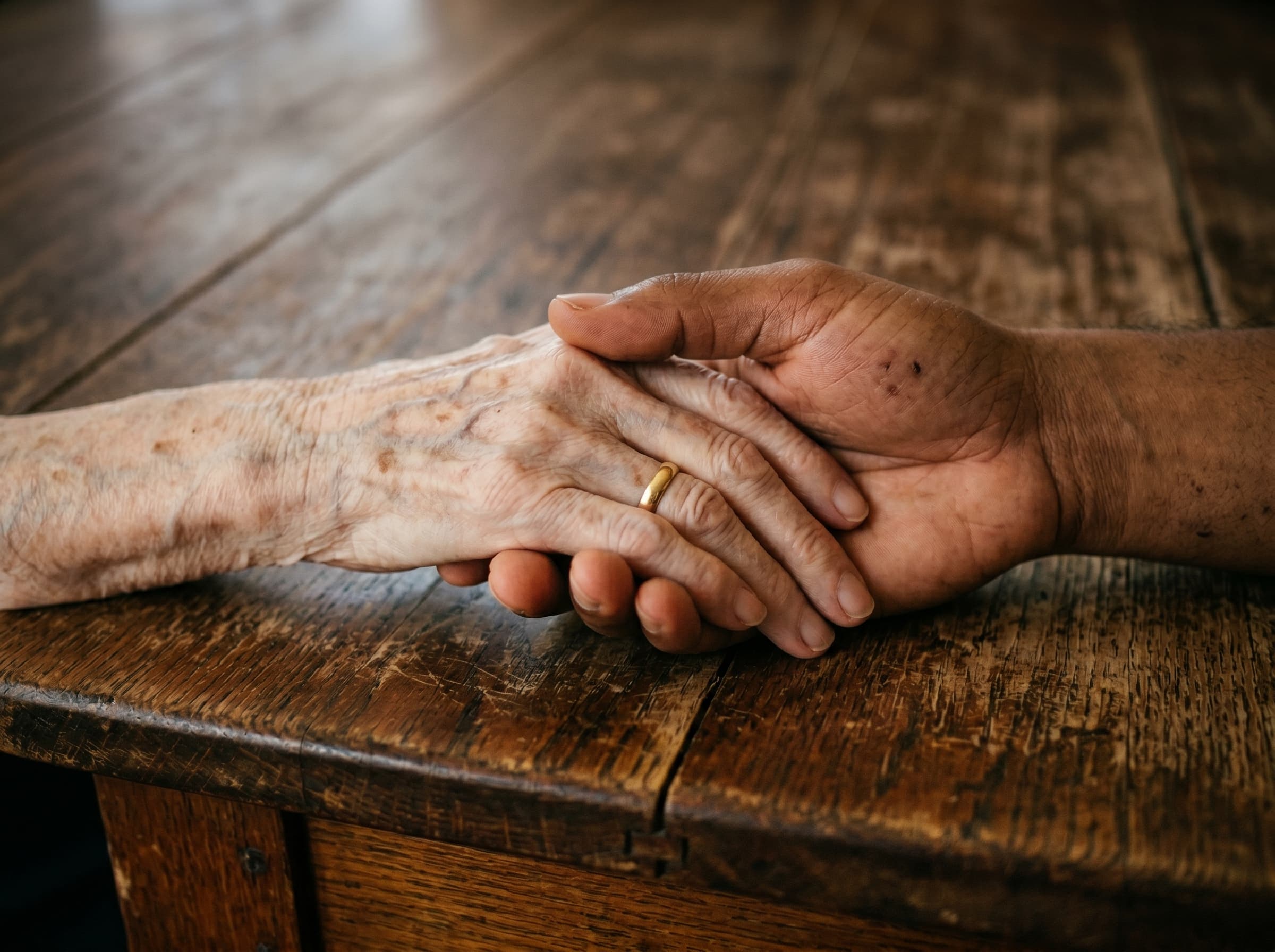 Two hands resting across a worn wooden kitchen table — one elderly, one younger.
